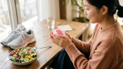 A woman's hands gently holding a pink ribbon near a healthy salad bowl and running shoes, symbolizing breast care and wellness.