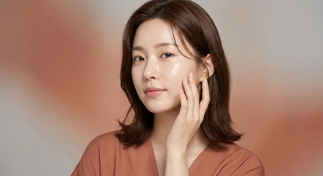 A beautiful young woman with flawless glowing skin gently touching her cheek, set against a warm terracotta background.